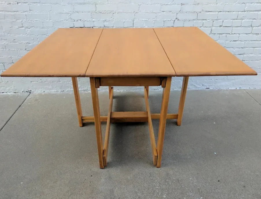 A mid-century modern Conant Ball solid maple drop-leaf table with a visible wood grain, set against a white brick background.